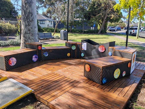 A photo of an outdoor parklet (raised area where people can sit in a park). The parklet is timber with blue and black panels. On the panels are artworks and colourful mosaics.
