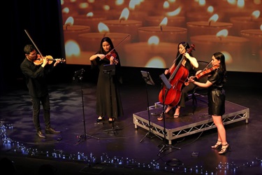 A string quartet plays on a stage. The backdrop is a full-screen picture of rows of glowing candles.