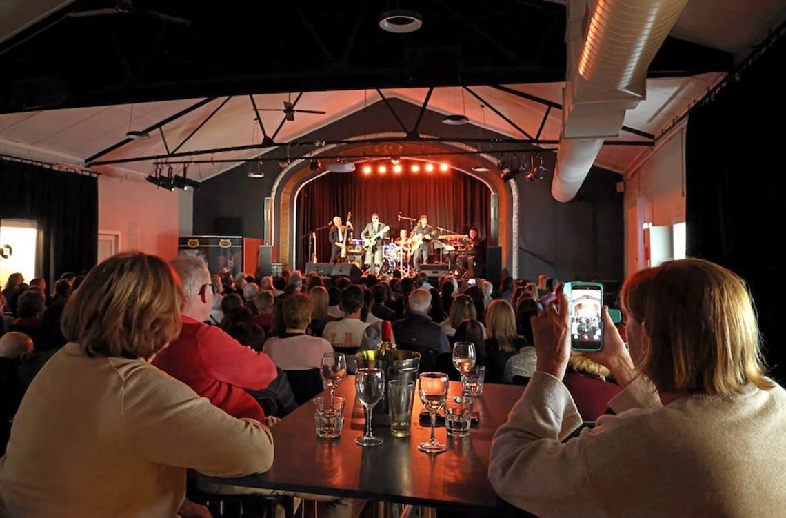 Long shot of a stage. Four people are on stage: an Elvis impersonator, two guitarists and a drum player. The crows is watching and sitting at tables filled with wine glasses. Some are filming on their phone.