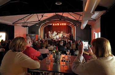 Long shot of a stage. Four people are on stage: an Elvis impersonator, two guitarists and a drum player. The crows is watching and sitting at tables filled with wine glasses. Some are filming on their phone.