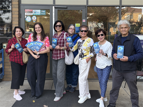 A group of 7 people proudly holding up Neighbourhood House Scavenger Hunt flyers for Neighbourhood House Week 2026